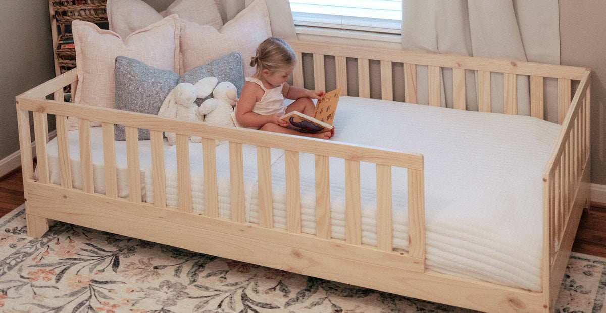 A toddler sits on a light natural wood Montessori style toddler floor bed with a white mattress, surrounded by beige and blue pillows, in a softly lit bedroom with curtains and a floral rug.
