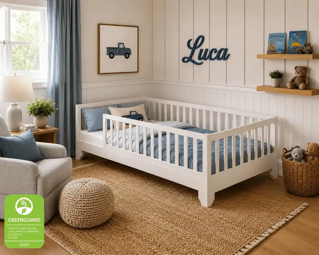 A white wooden toddler floor bed with a guard rail, styled in a bright, modern children's bedroom with blue bedding, a woven rug, and a green Greenguard Gold badge in the bottom-left corner.