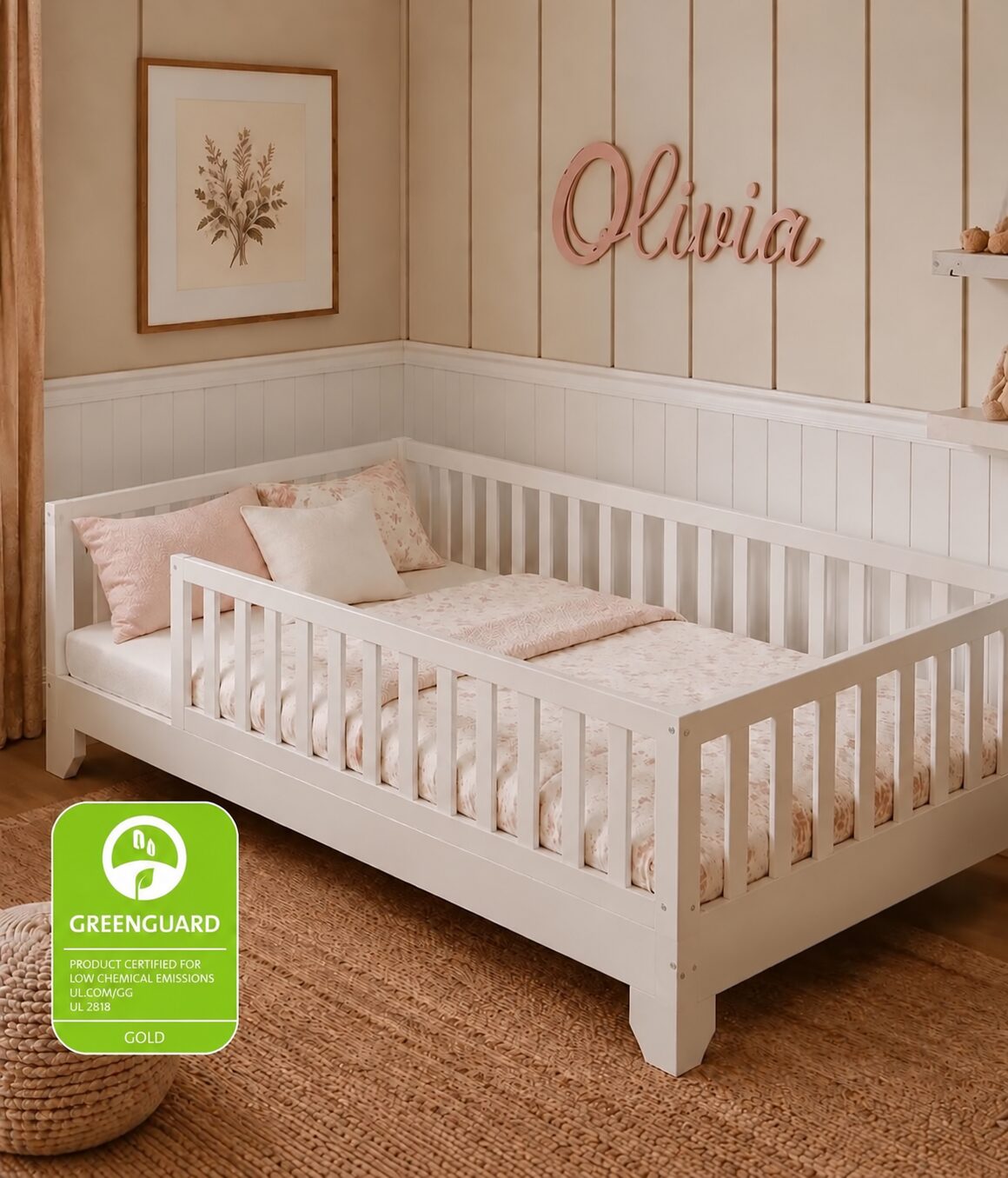 A white wooden toddler floor bed with a guard rail and light pink bedding, shown in a styled nursery setting with beige walls, a framed botanical print, and a wooden 'Olivia' wall sign. Text at the top announces a November delivery, and a green Greenguard certification badge appears in the bottom left.