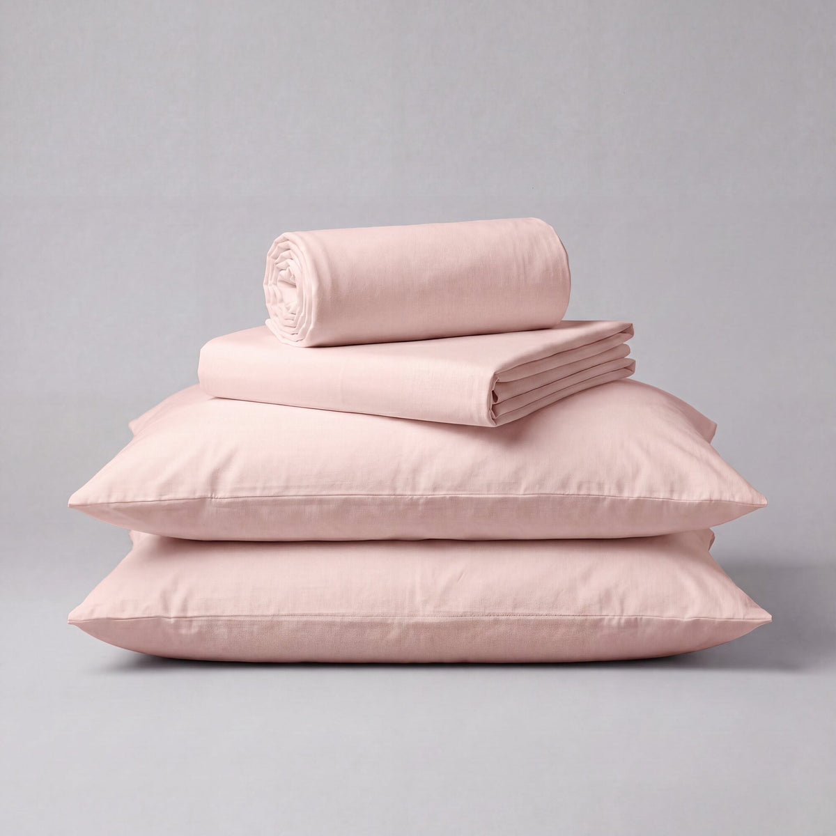 A stack of mauve organic toddler bedding, including two flat pillowcases, a folded sheet, and a rolled top layer, displayed on a neutral gray background.