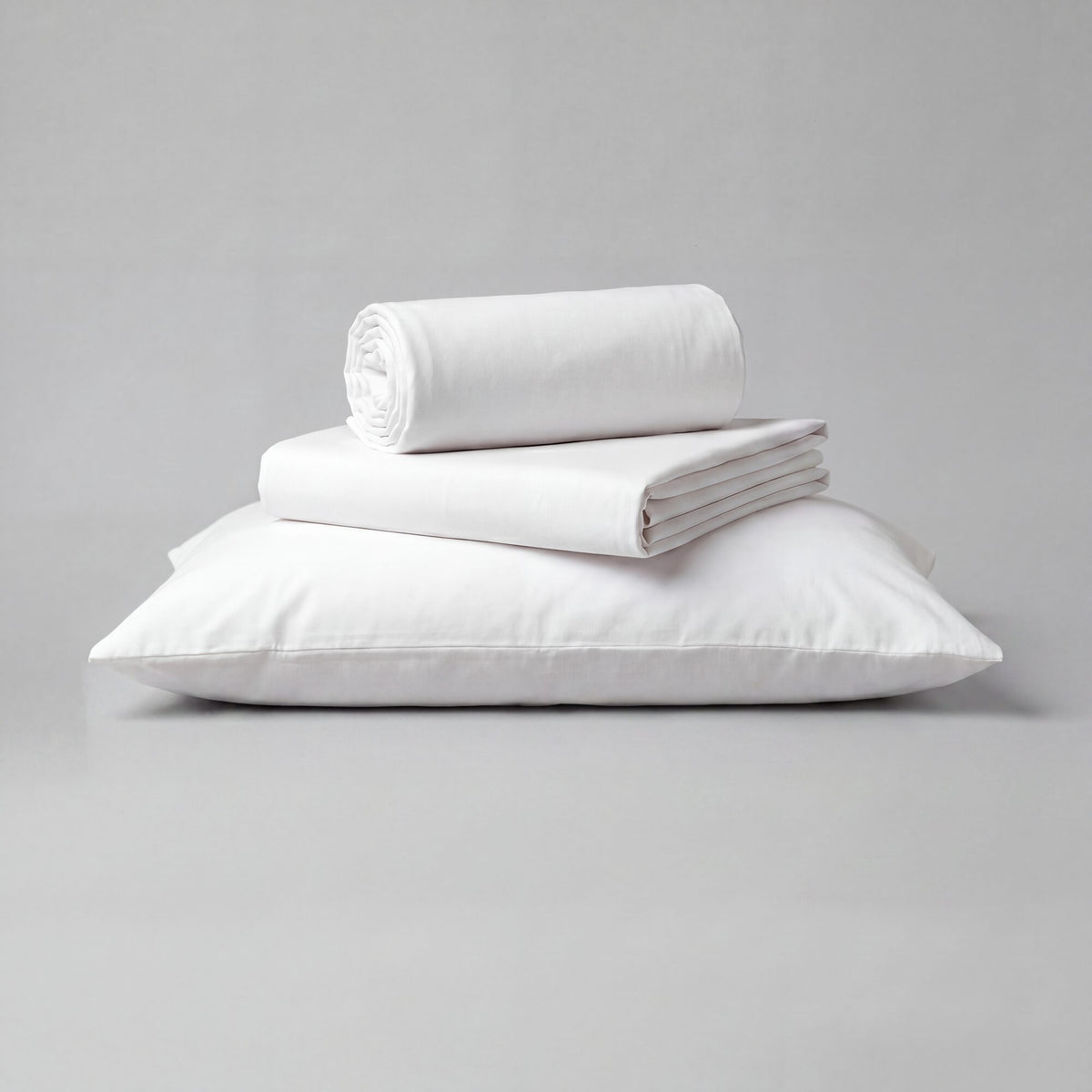 A stack of white toddler sheets, including a folded pillowcase, a folded sheet, and a rolled sheet, displayed on a light gray background.