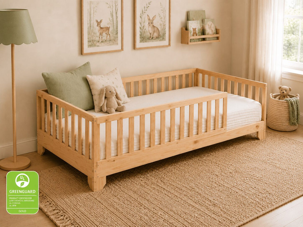 A light natural wood daybed with safety rails, styled in a warm, neutral-toned children's room with a green lamp, framed deer prints, and a woven rug. A green 'Greenguard Gold' certification badge is visible in the bottom-left corner.