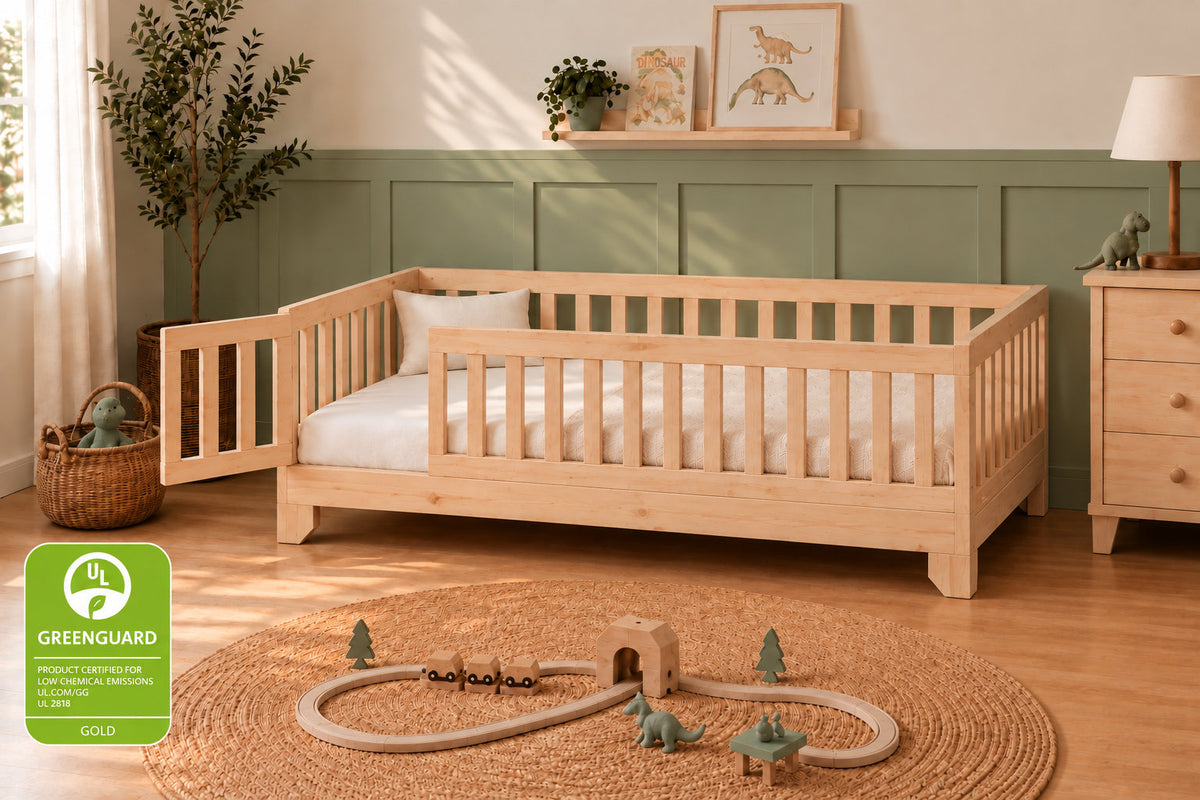 A light natural wood toddler floor bed with built-in side rails, styled in a Montessori child's bedroom with green paneled walls, a woven rug, dinosaur toys, and a green UL GreenGuard Gold badge in the lower-left corner.