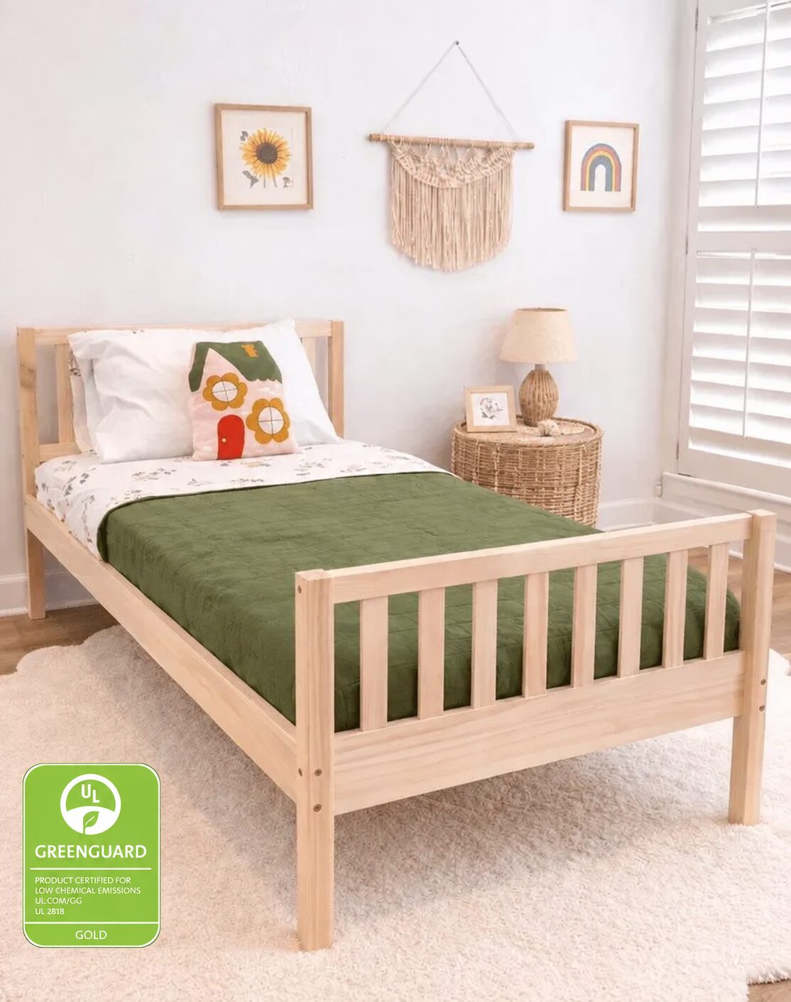 A light natural wood single bed frame with a green mattress cover and white bedding, styled in a bright children's bedroom on a scalloped white shag rug. A green GREENGUARD Gold certification badge is in the bottom-left corner.