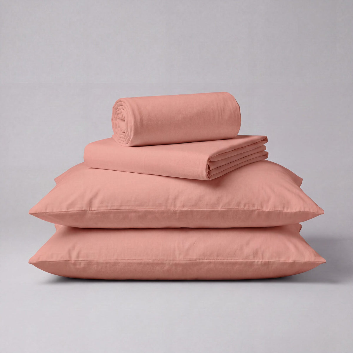 A stack of dusty pink bedding, including two flat pillowcases, a folded sheet, and a rolled duvet cover, displayed on a neutral light gray background.